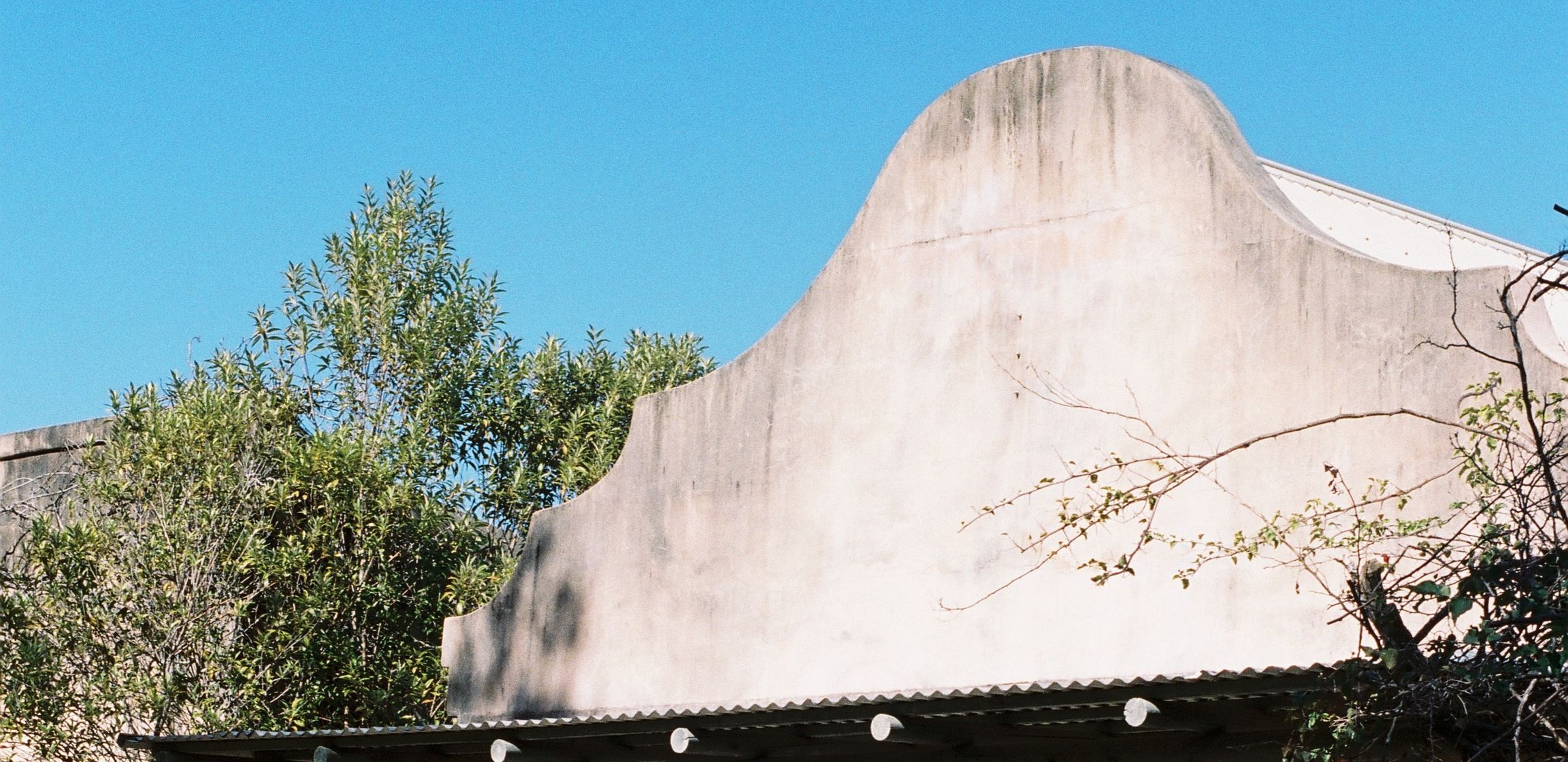 The front of Chalet Byron: a curved rendered parapet over a timber verandah, trees and blue sky